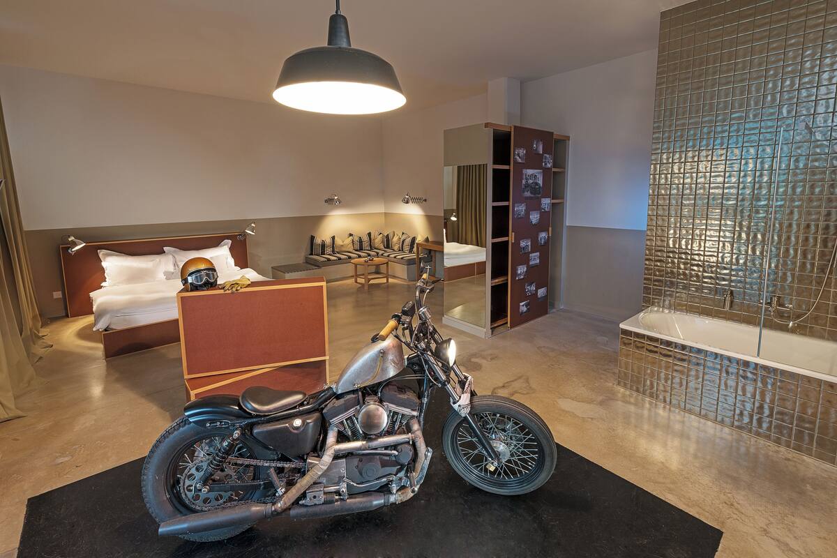 Habitación de hotel moderna con cama, zona de estar, una motocicleta y un baño con pared de azulejos.