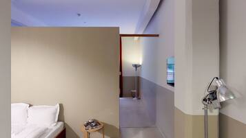 A hotel room with a bed, a small table with a magazine, a lamp, and a hallway leading to a bathroom.