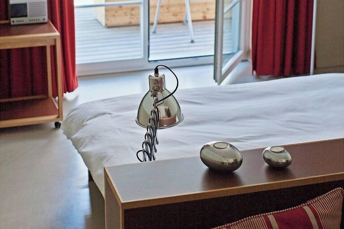 Habitación de hotel con cama, mesita de noche con lámpara, radio y vista a la playa a través de un ventanal grande.
