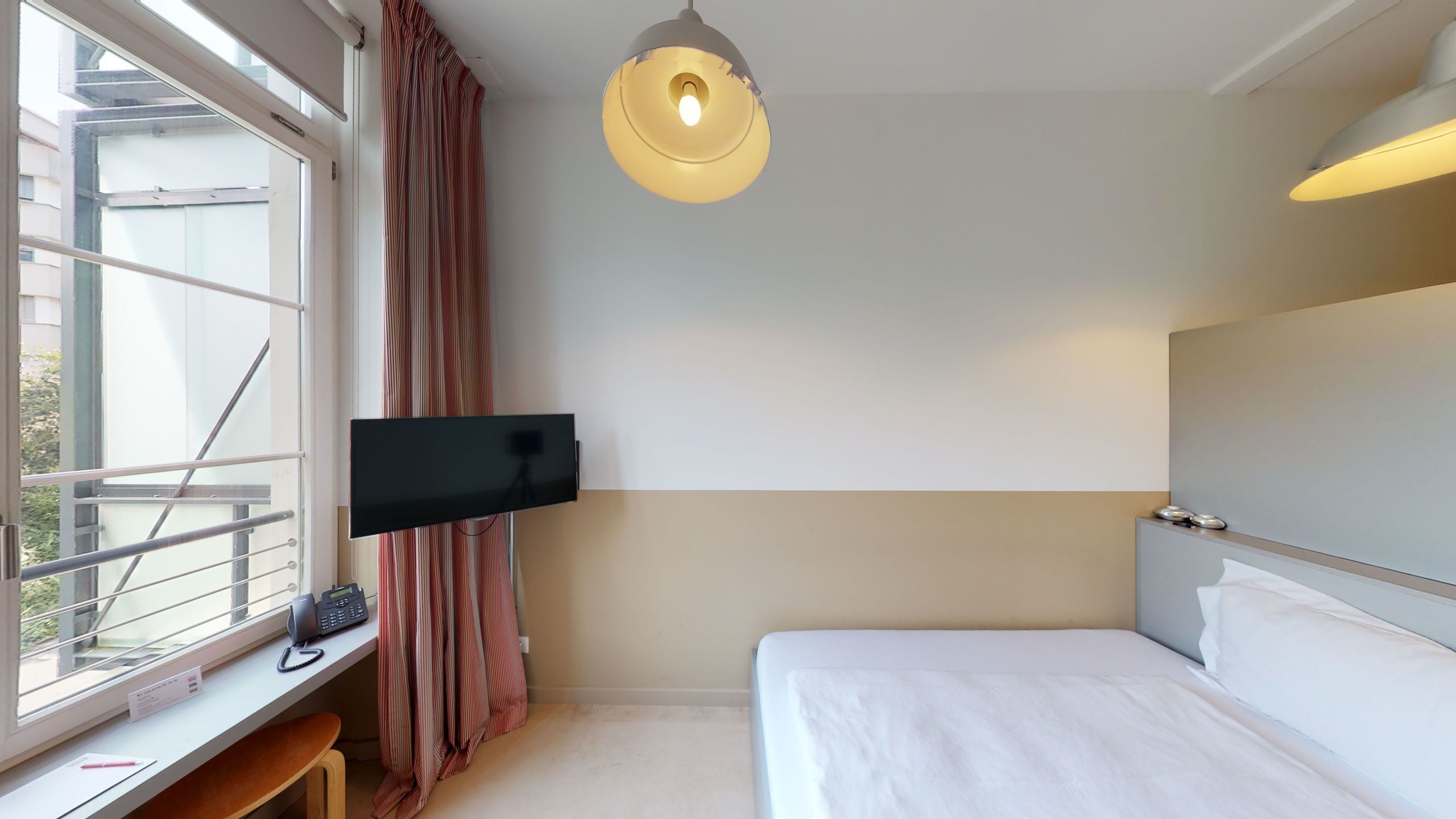 Hotelkamer met een bed, een bureau met computer, een aan de muur bevestigde televisie en een raam met gordijnen.