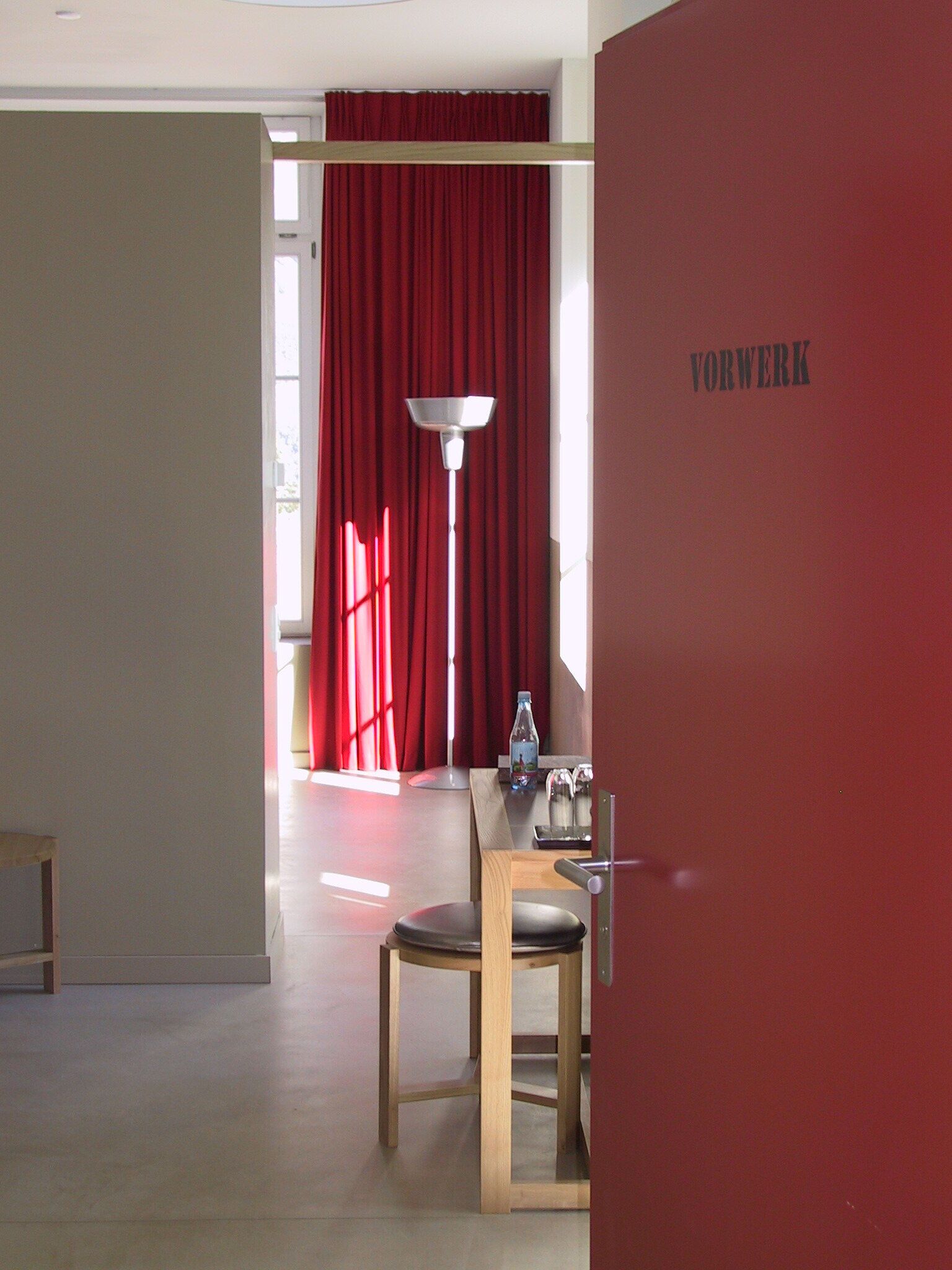 Een kamer met een eettafel, een stoel, een lamp en een rode muur waarop 'VORWERK' staat.