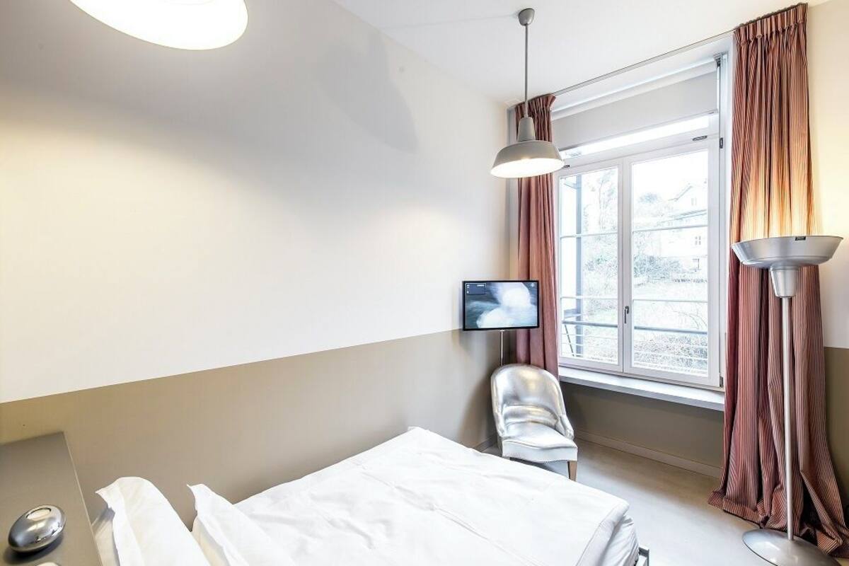 Habitación de hotel con cama, una silla, televisor, ventana con cortinas y una lámpara.