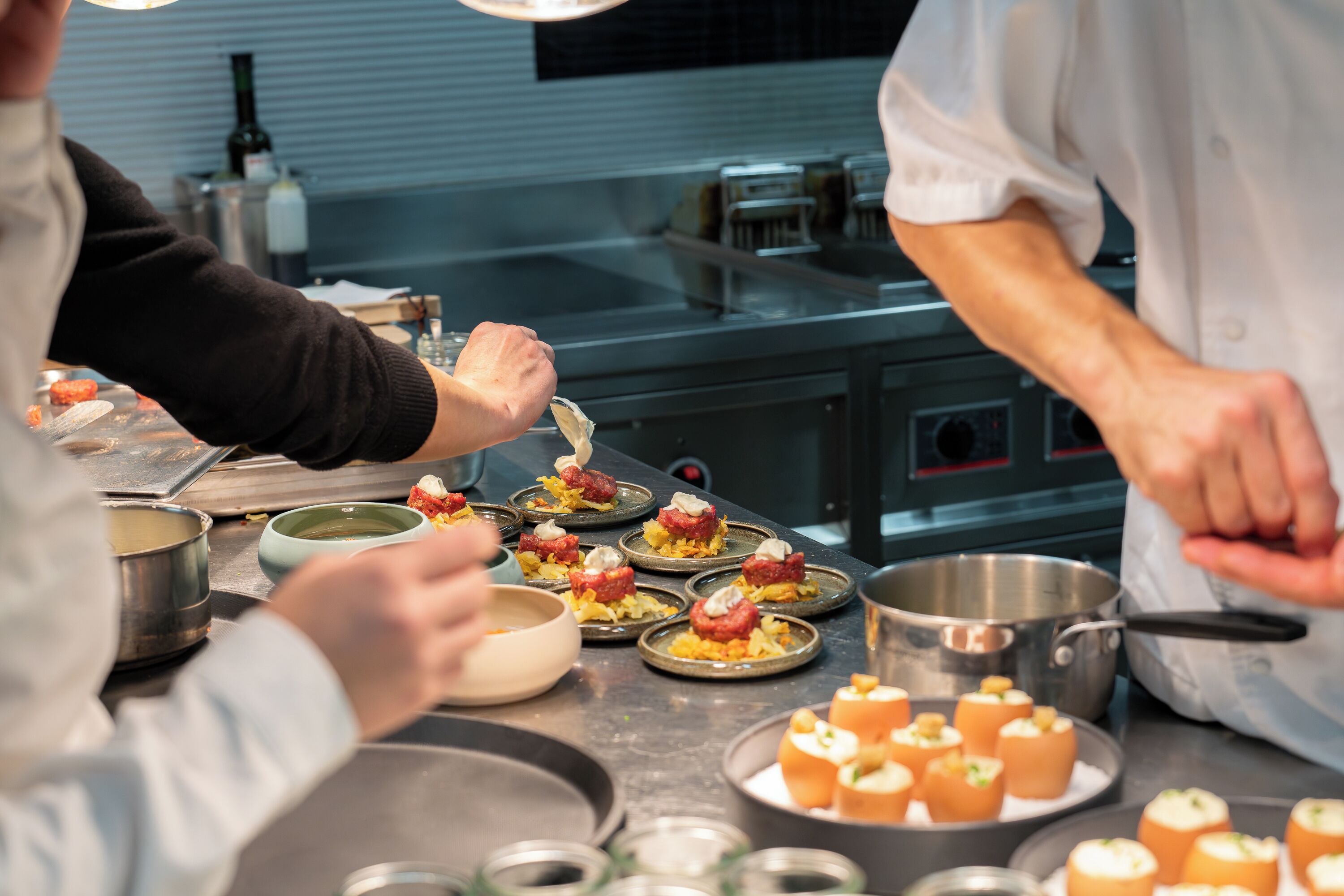 Una cocina con chefs preparando alimentos, electrodomésticos de acero inoxidable y una variedad de platos en la encimera.
