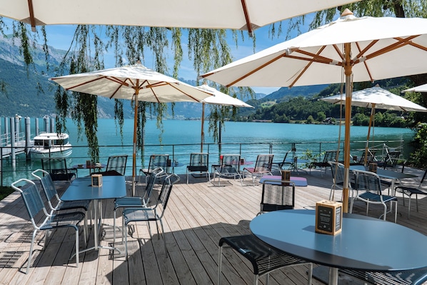 Eine Terrasse mit Sitzgelegenheiten, Sonnenschirmen und Blick auf einen See und Berge.