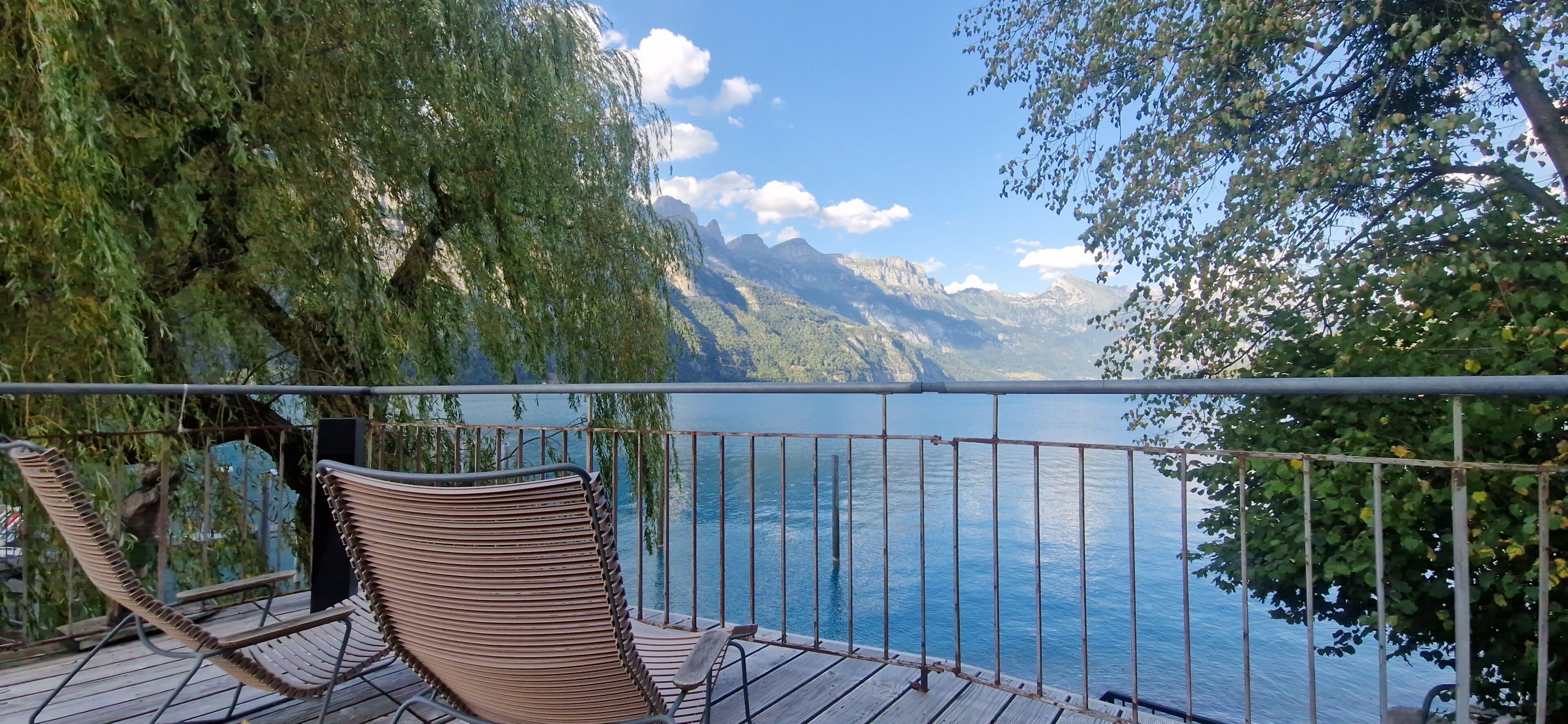 Una terraza de madera con barandas metálicas da a un lago tranquilo, rodeado de vegetación exuberante y montañas a lo lejos.