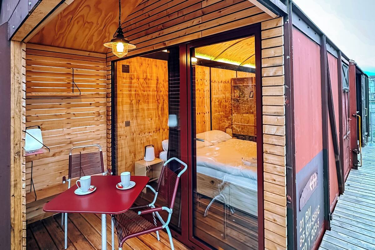 Una cabaña acogedora con interior de madera, un comedor con mesa y sillas, y una cama visible a través de un vidrio.