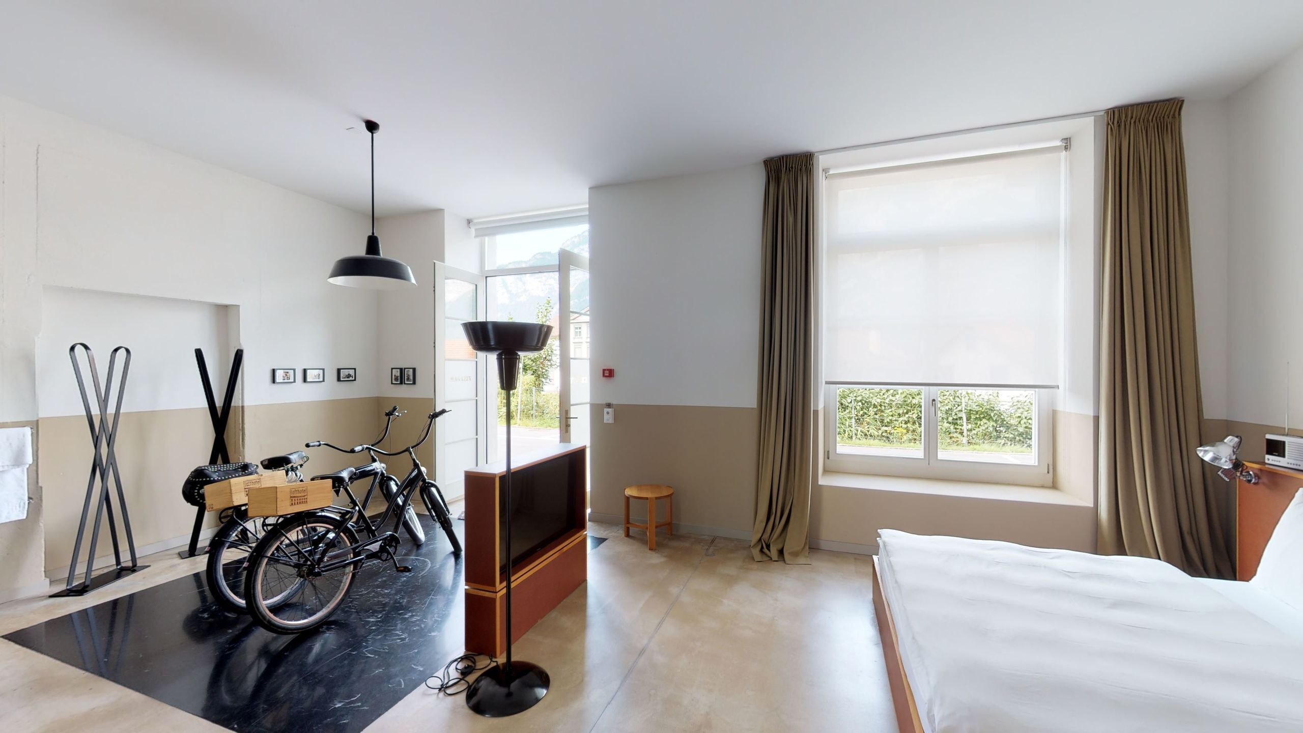 A hotel room with a bed, a desk, a bicycle, a chair, a lamp, and a window with curtains.