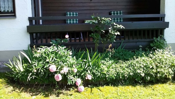 A building with a wooden balcony, a window with a curtain, and a garden with pink flowers. - Ferienwohnung, 35 qm Erdgeschoss, Kombinierter Wohn- und Schlafraum (Bad Endorf)