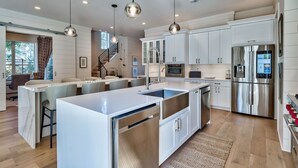Una cocina moderna con armarios blancos, electrodomésticos de acero inoxidable, una isla central, iluminación colgante y una escalera que lleva a un comedor.