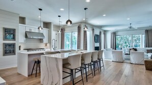 Una cocina moderna con una isla de mármol, iluminación colgante y un comedor con mobiliario blanco.