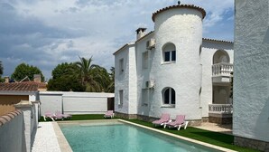 Uma área de piscina com um edifício branco, piscina azul cristalina e espreguiçadeiras listradas em vermelho e branco.