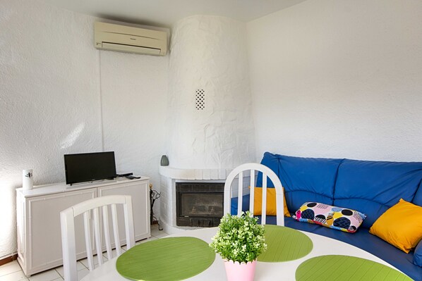 Un salon avec une cheminée blanche, un canapé bleu agrémenté de coussins colorés, un bureau blanc avec un ordinateur et une plante verte posée sur une table.