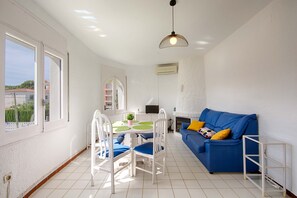 A bright living room with a blue sofa, white chairs, a dining table with green placemats, and a TV. - BADIA 4,PISO 2ha,PISCINA,100m PLAYA,PARKING,WIFI (Empuriabrava)