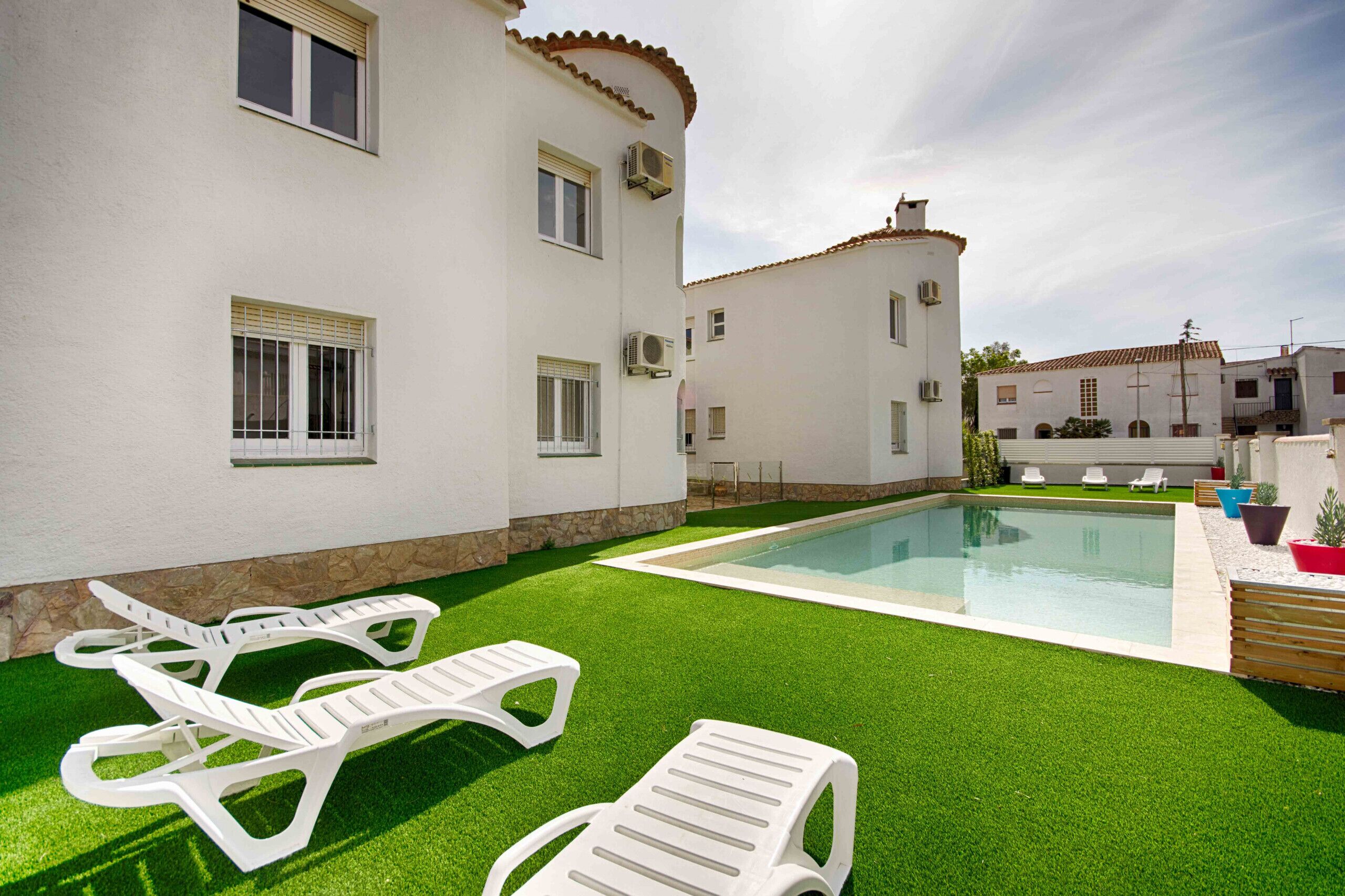 Una piscina con césped artificial, sillas de descanso blancas y un edificio blanco con varias ventanas y unidades de aire acondicionado.