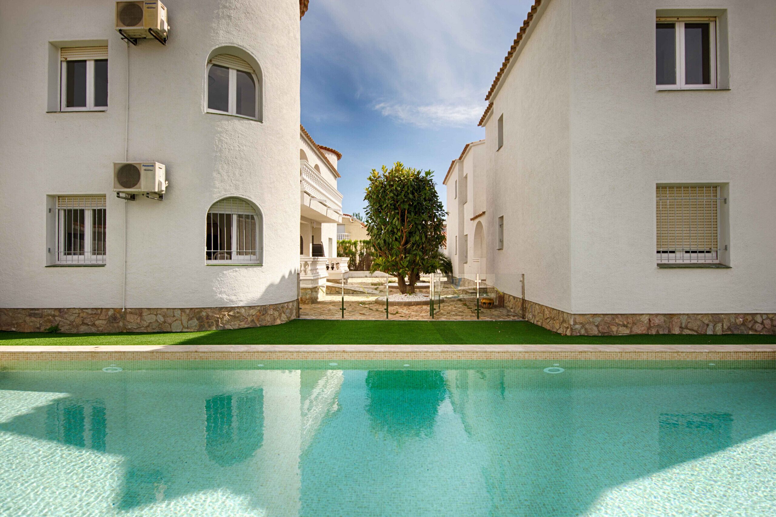 A pool area with a clear view of the pool, surrounded by white buildings with arched windows and a tree in the center.
