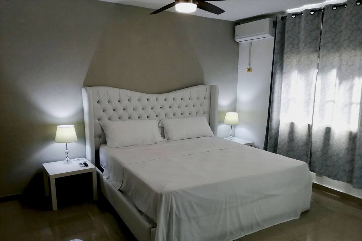 A neatly made bed with a tufted headboard, two bedside tables with lamps, a ceiling fan, and a window with curtains.