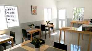 A dining area with wooden tables and chairs, a small kitchen area with a sink, and a window with a view of greenery. - Hotel Yamir (Barahona)