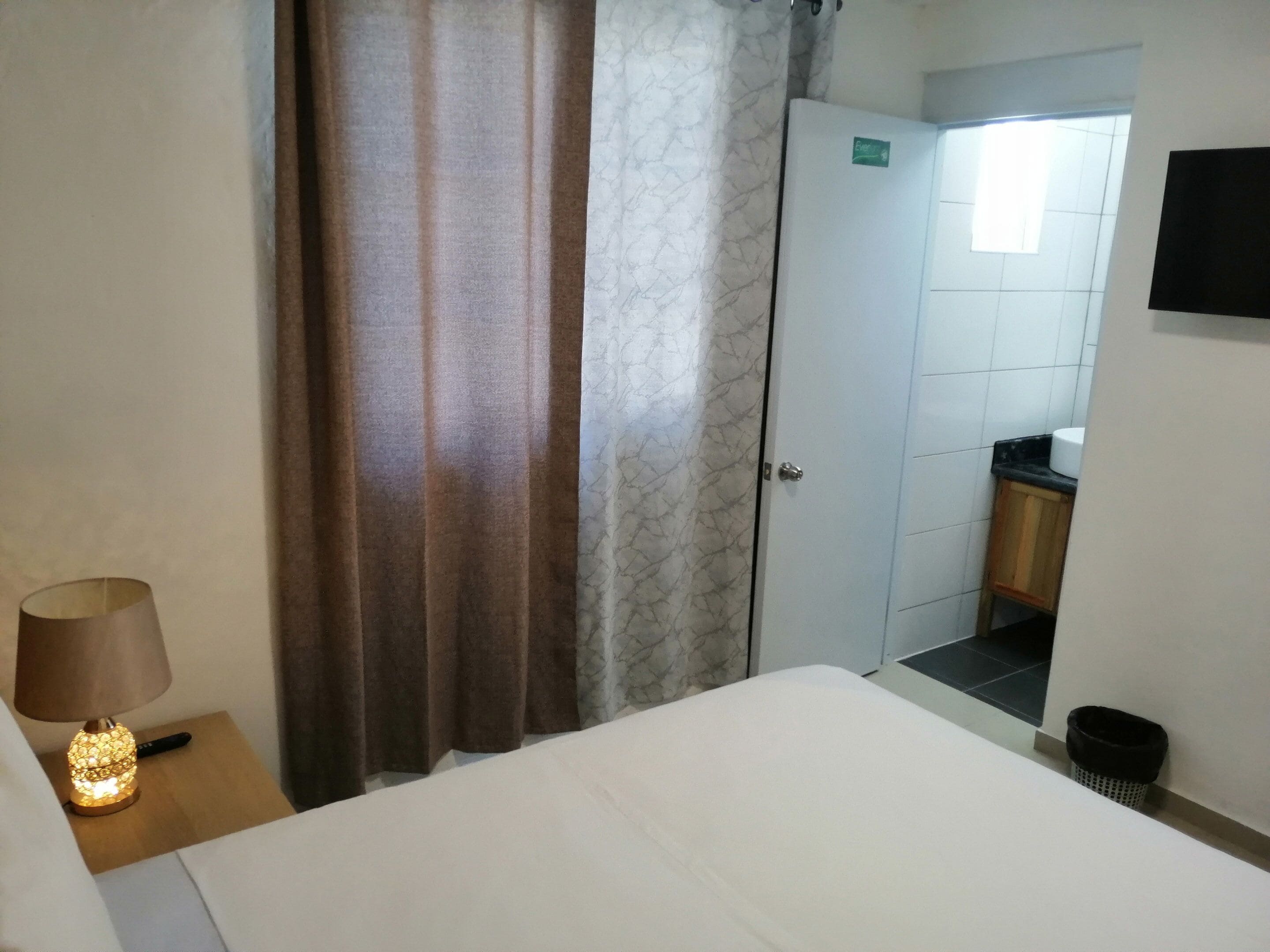 A hotel room with a bed, a nightstand with a lamp, a bathroom with a toilet and sink, and a door leading to another room.
