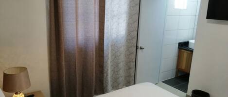 A hotel room with a bed, a nightstand with a lamp, a bathroom with a toilet and sink, and a door leading to another room.