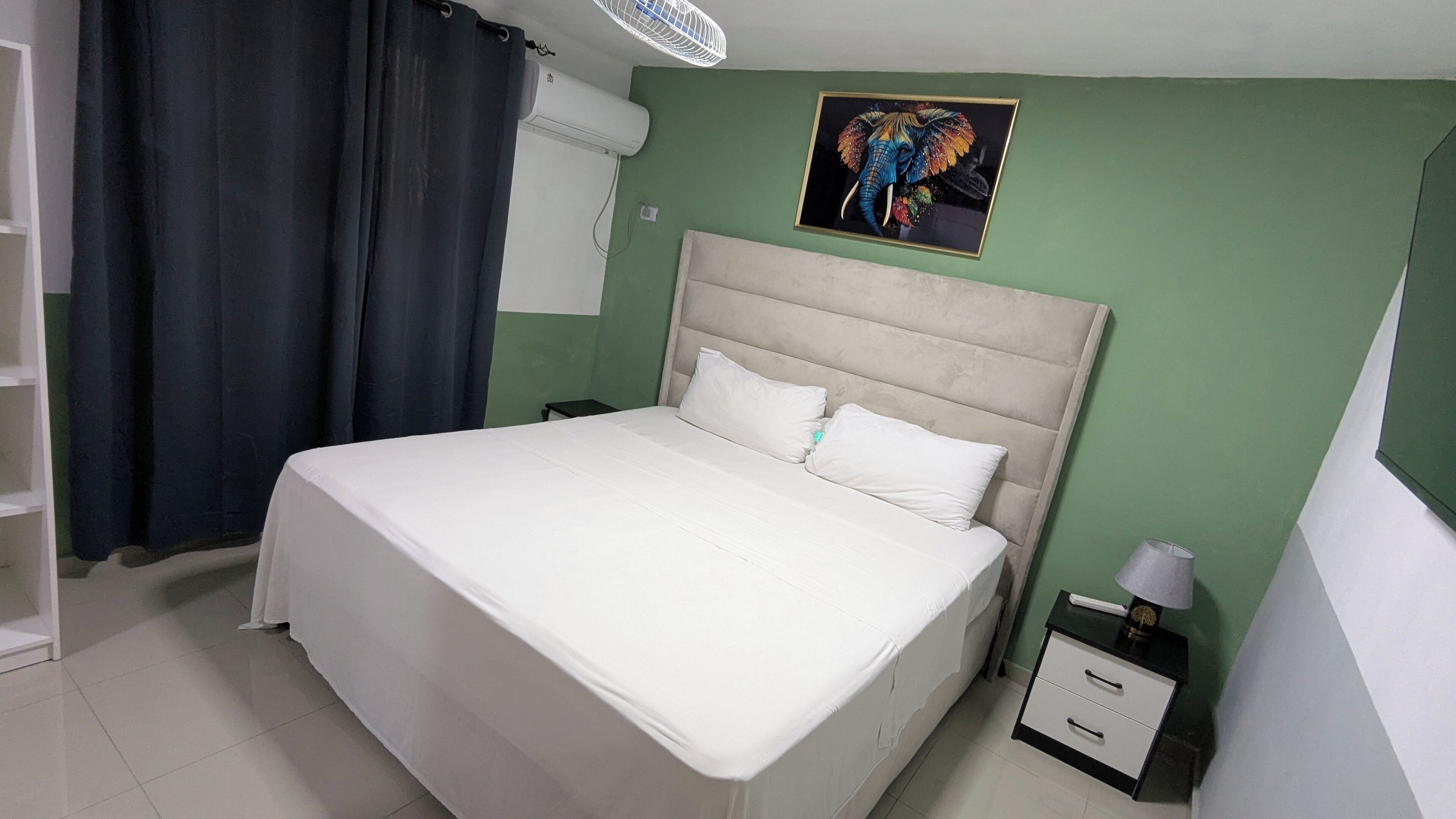 A neatly made bed with white linens in a room with green walls, a white ceiling fan, and a framed picture of an elephant.