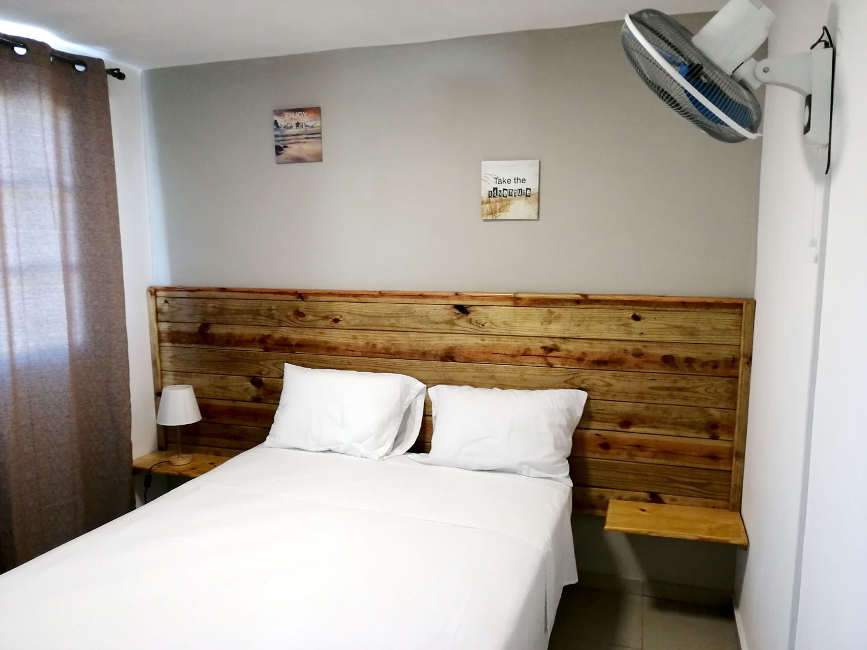 Une chambre à coucher avec une tête de lit en bois, deux oreillers blancs, une lampe de chevet et un ventilateur fixé au mur.