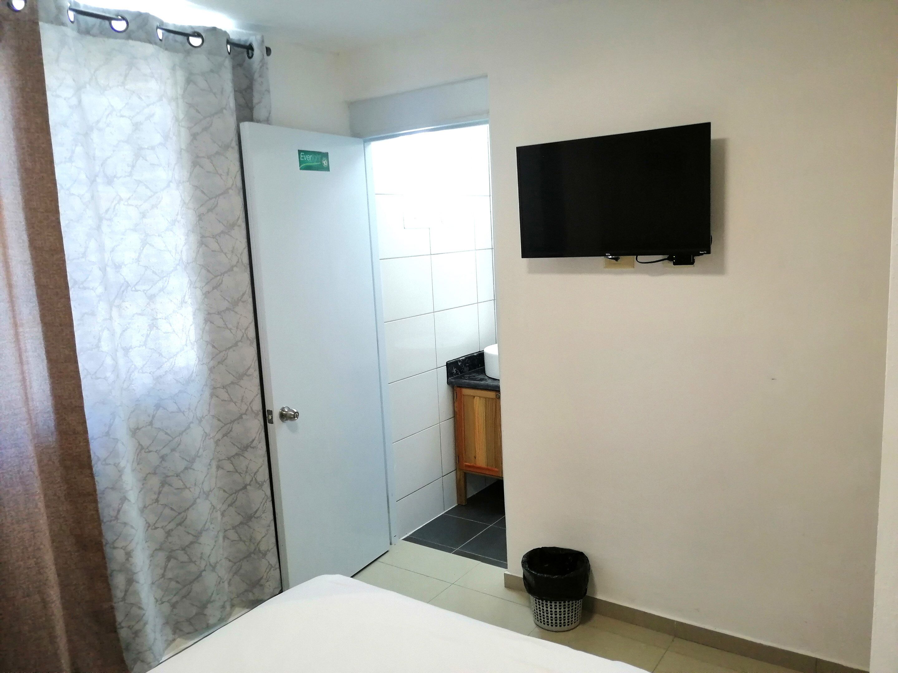 A hotel room with a bed, a television mounted on the wall, a bathroom with a toilet and sink, and a shower curtain.
