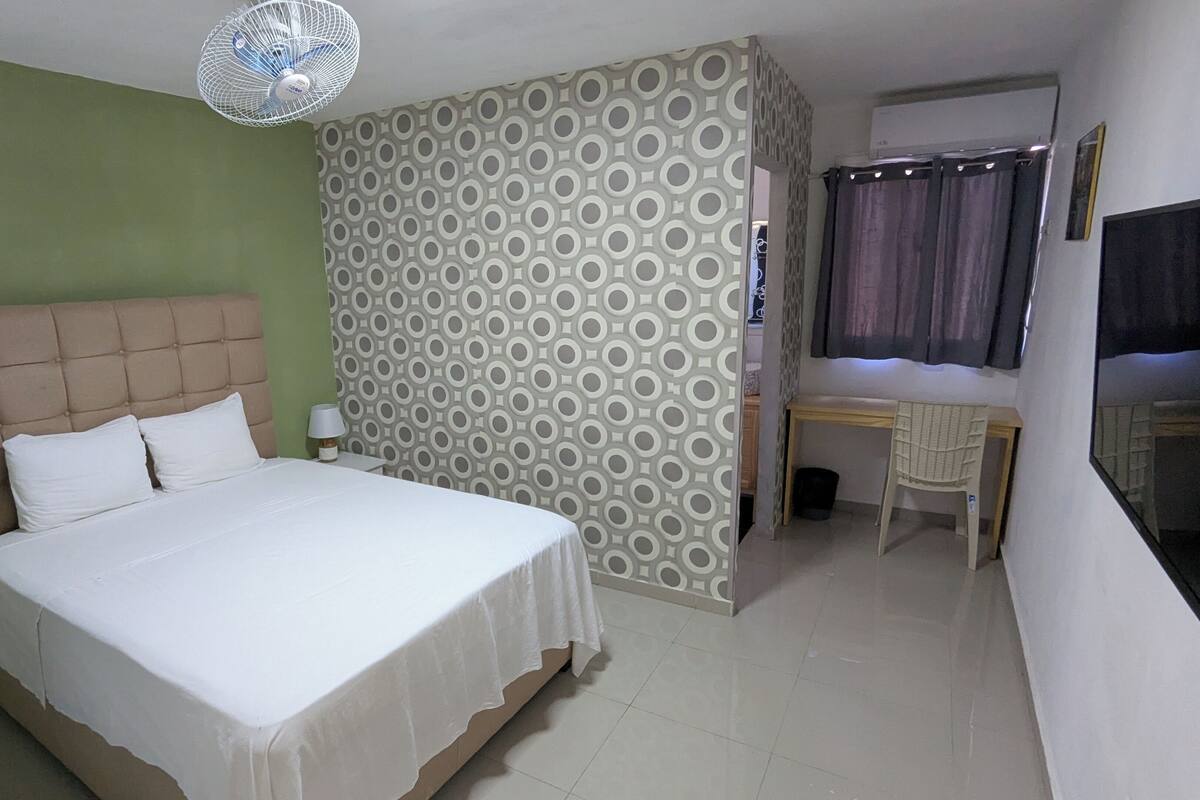A hotel room with a bed, a desk, a chair, a standing fan, and a patterned wall.