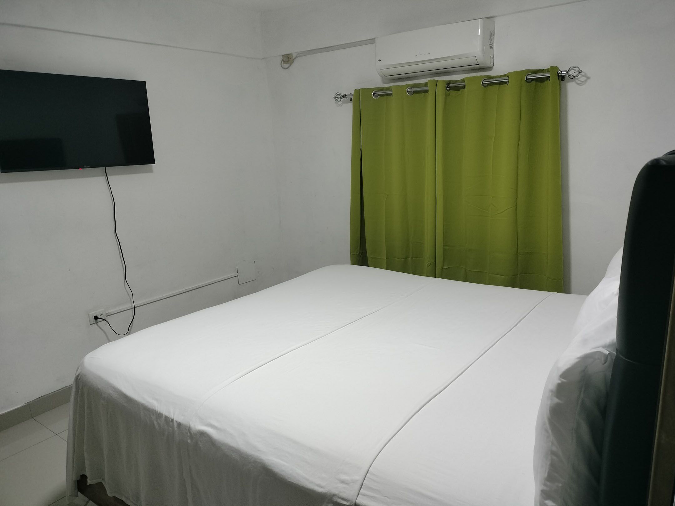 A single bed with white bedding, a flat-screen TV mounted on the wall, and a green curtain.