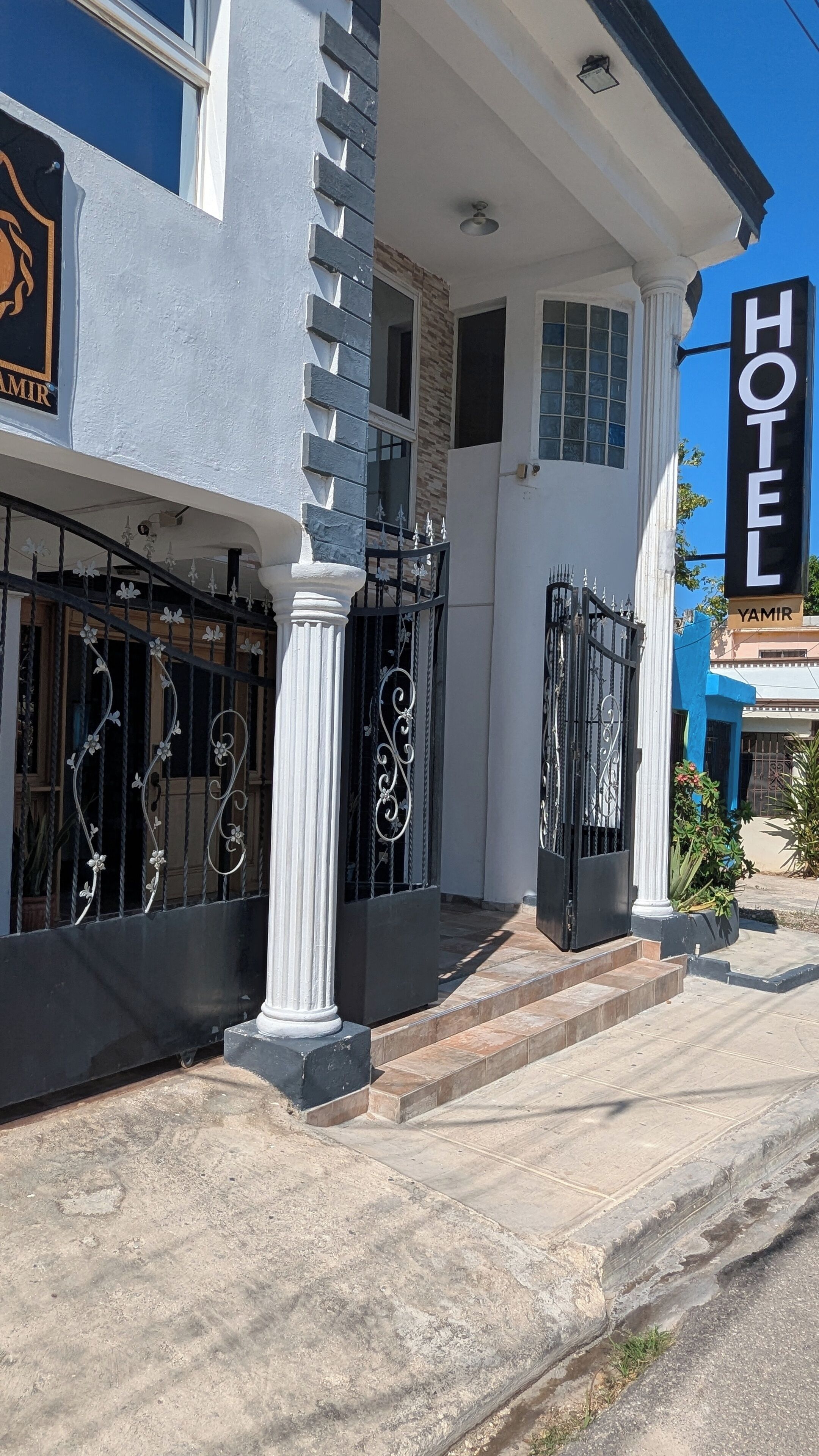 Entrada de un hotel con una verja negra, columnas blancas y un cielo azul despejado.