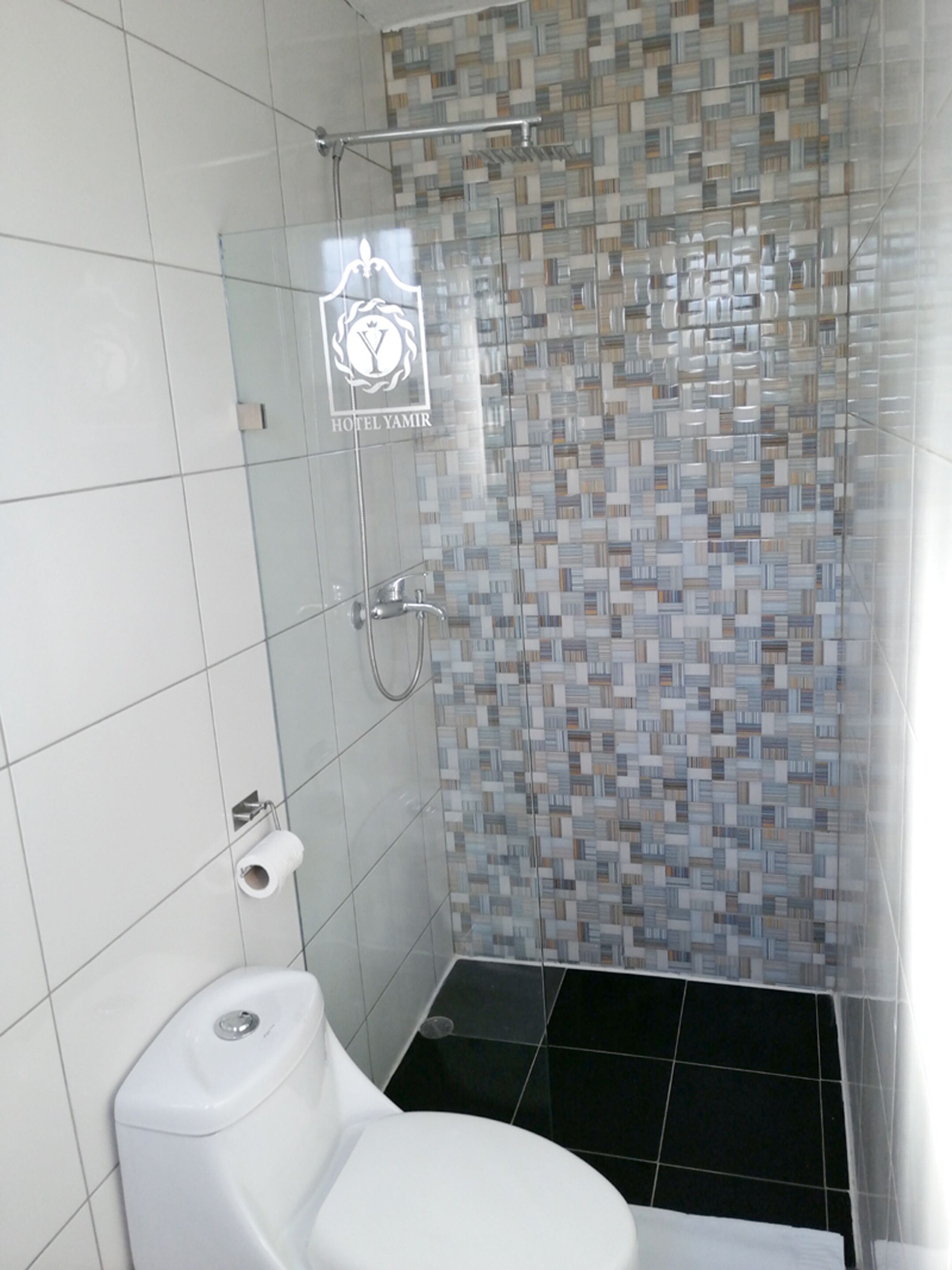 Un baño con inodoro, ducha con puerta de vidrio y pared revestida de azulejos con el logotipo del hotel.