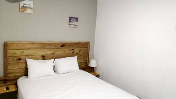 Un dormitorio con cabecera de madera, dos almohadas blancas, una colcha blanca, una lámpara de noche y dos letreros en la pared.