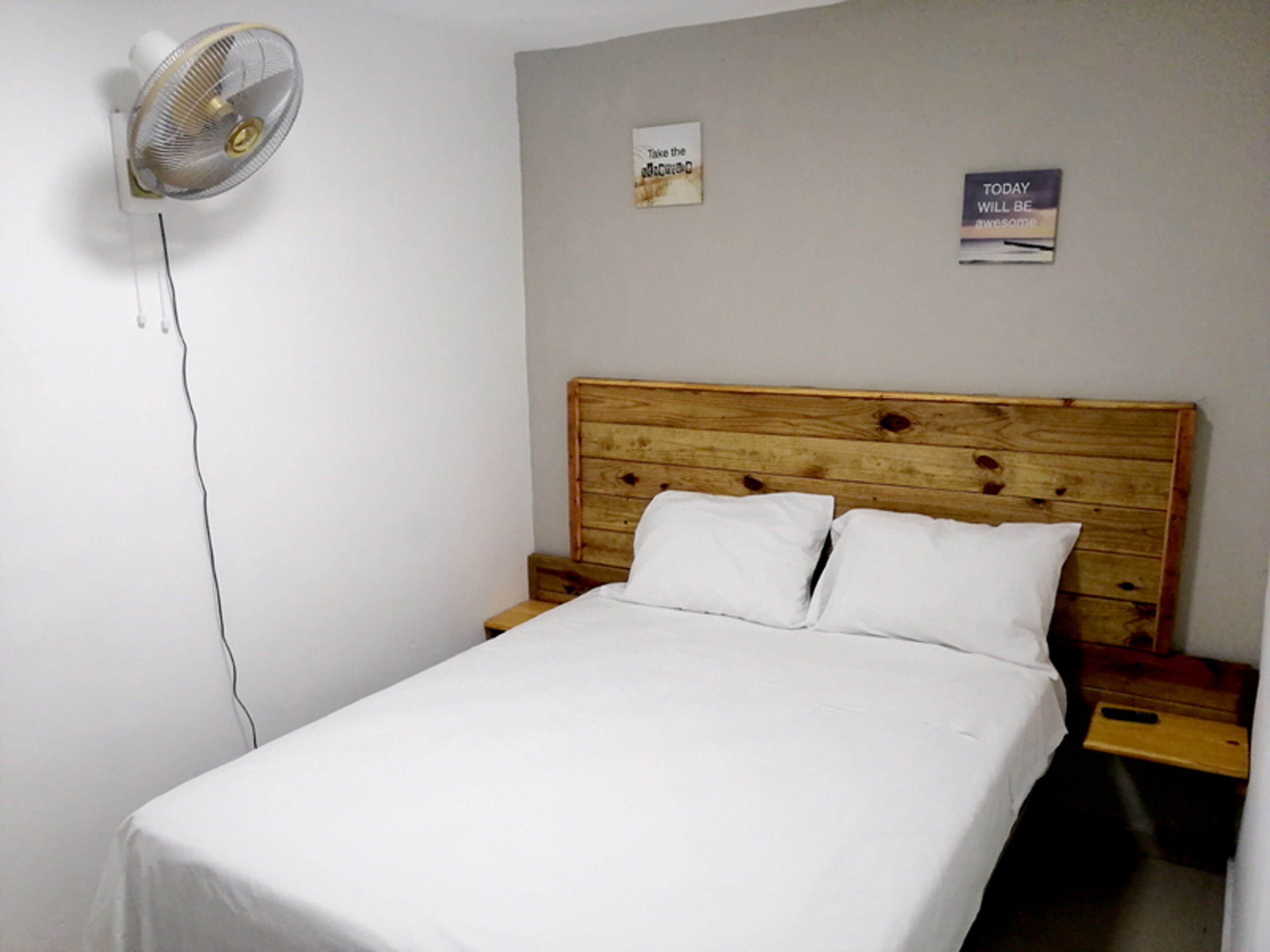 Una cama individual con cabecera de madera, ropa de cama blanca y un ventilador fijado a la pared.