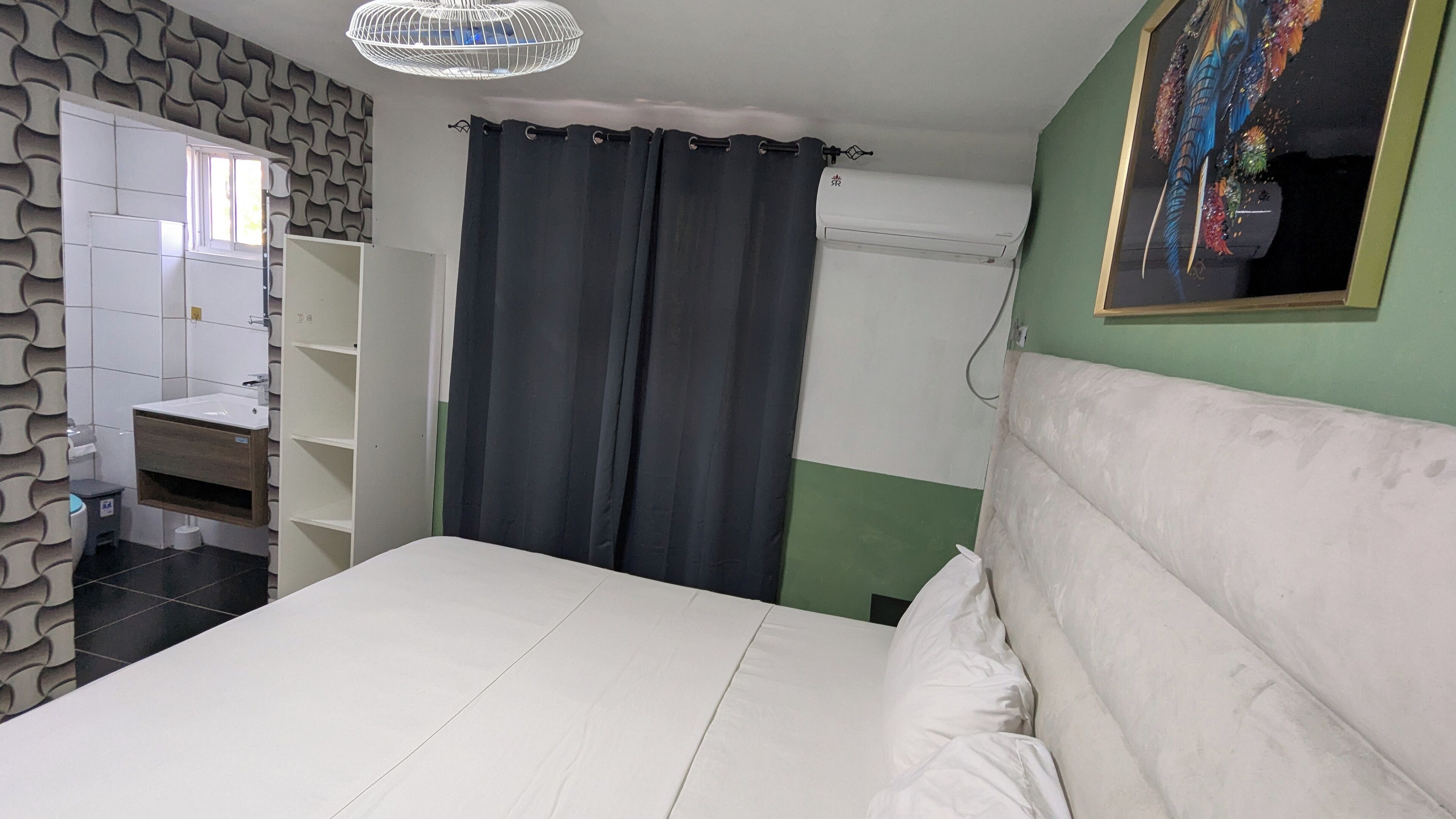 Ein Schlafzimmer mit einem Bett, einem weißen Kopfteil, einem Nachttisch, einem Spiegel, einem Fenster mit Vorhängen, einem Kleiderschrank und einer wandmontierten Klimaanlage.
