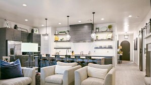 A modern kitchen with dark cabinetry, pendant lighting, and a central island. There are two armchairs and a sofa in the living area.