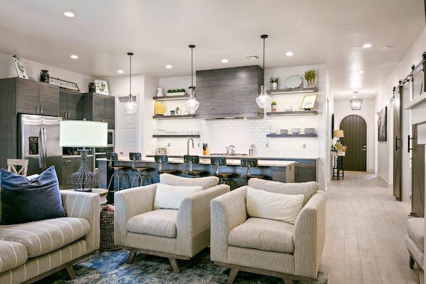 Een moderne keuken met donkere kastjes, hanglampen en een centraal eiland. In de woonkamer staan twee fauteuils en een bank.
