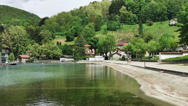 Um lago tranquilo com um pequeno pato amarelo flutuando, cercado por colinas verdes exuberantes e alguns edifícios ao fundo.