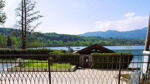 Terraço com duas cadeiras estofadas verdes, uma mesa com um livro e um vaso de planta, e vista para um lago e montanhas.