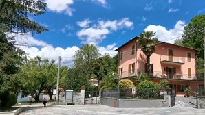 Um edifício de vários andares com varandas, uma área pavimentada em frente e um corpo de água ao fundo.