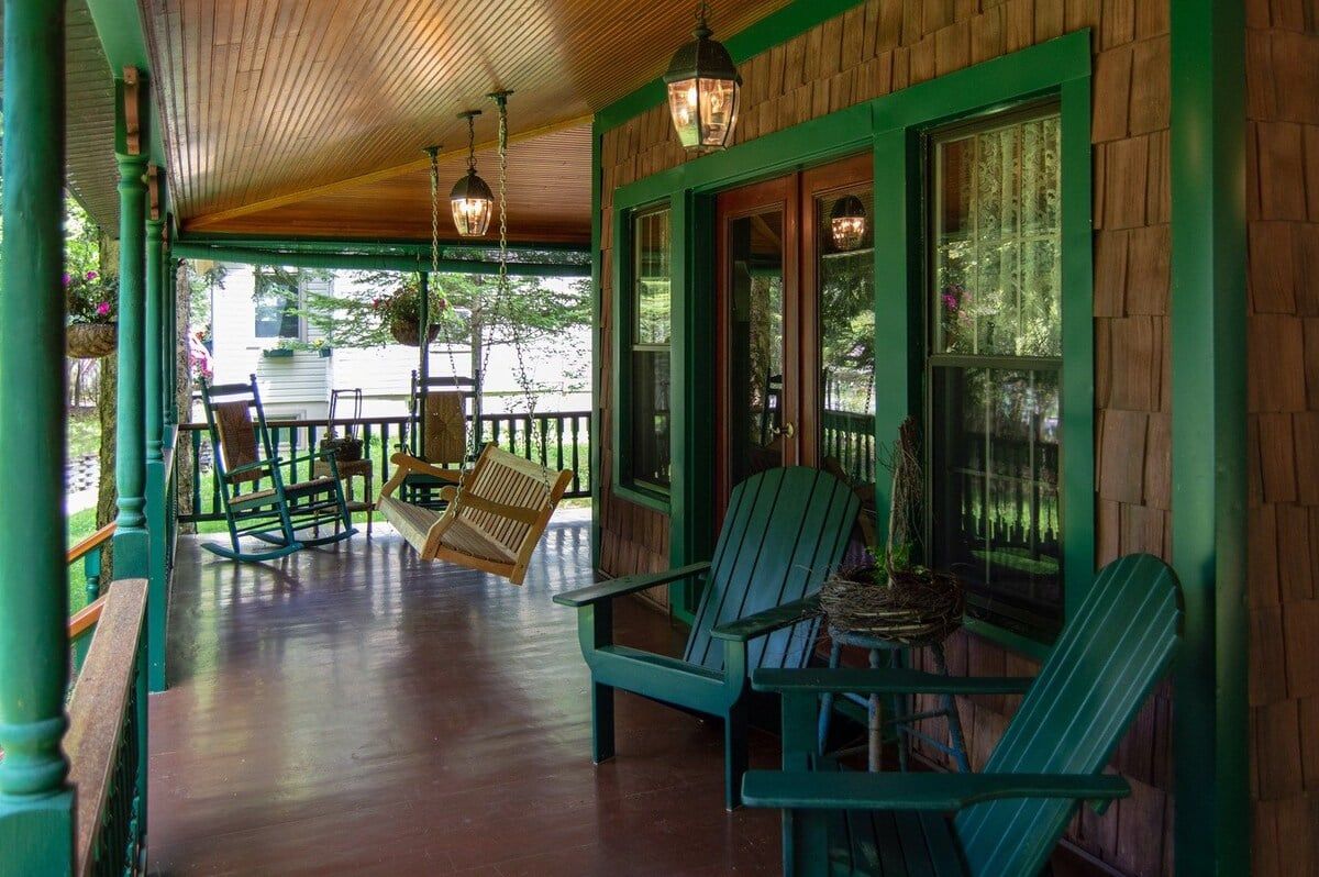 A covered porch with green Adirondack chairs, a rocking chair, and hanging lanterns.