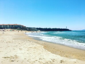Uma praia com areal, algumas pessoas e edifícios ao fundo.