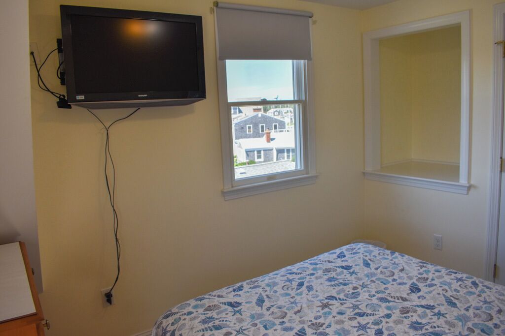 Ein Schlafzimmer mit einem Bett, einem an der Wand befestigten Fernseher, einem Fenster mit Blick auf Gebäude und einer Tür.
