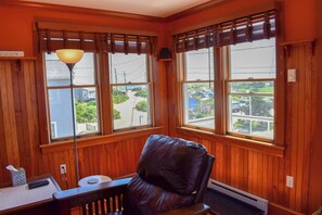 A room with wooden paneled walls, a leather chair, a desk with a lamp, and large windows with blinds.