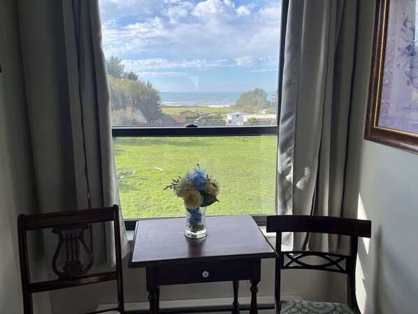 A room with a view of a grassy field and the ocean through a window, a small table with a vase of flowers, and two chairs.