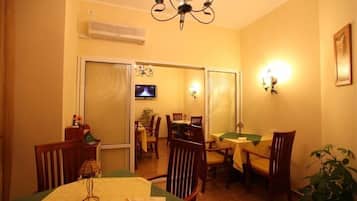 A dining area with wooden chairs and tables, a chandelier, and a television on the wall.