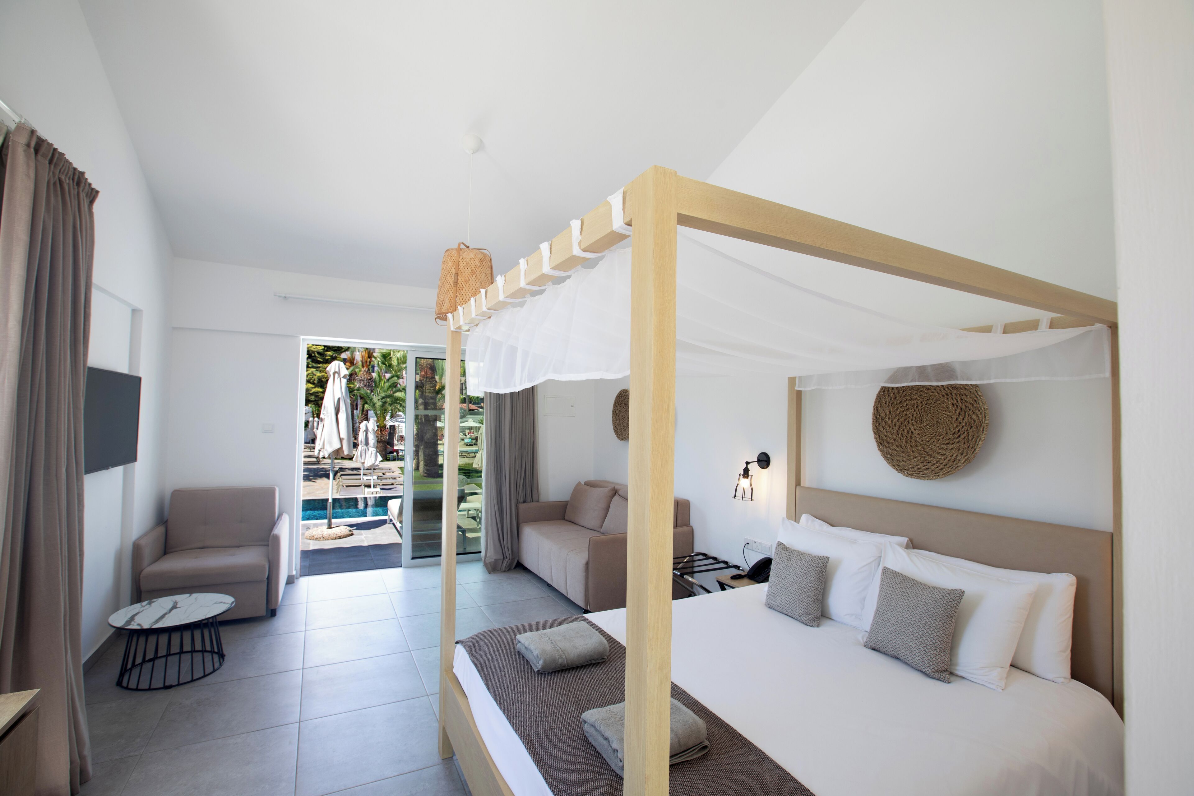 Ein modernes Hotelzimmer mit einem großen Bett, einer Couch, einem kleinen Tisch und Blick auf den Poolbereich.