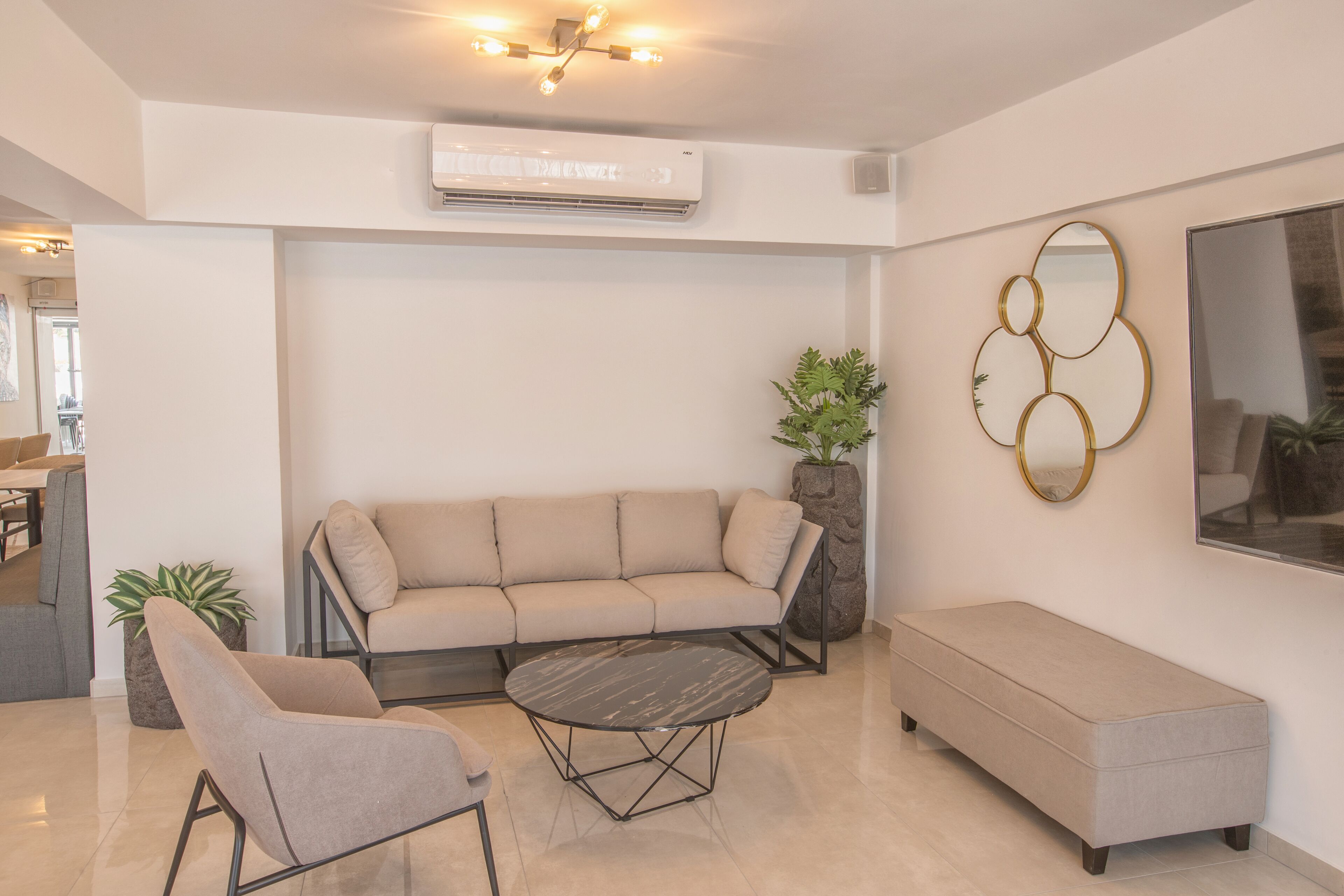 Een moderne woonkamer met een beige bank, een ronde salontafel en een bankje. Er staat een potplant en een decoratieve spiegel aan de muur.