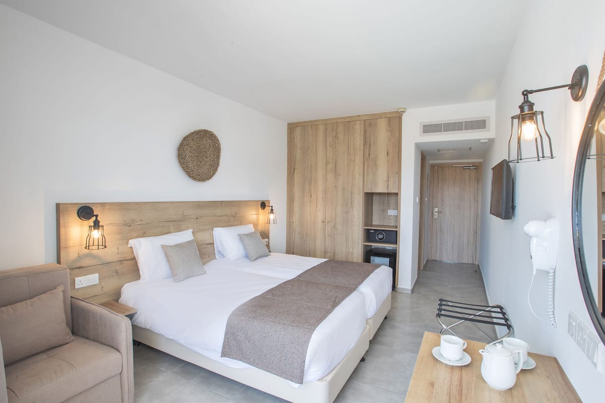 A modern bedroom with a large bed, bedside tables, a chair, a wooden wardrobe, a mirror, and a small table with cups and a pitcher.