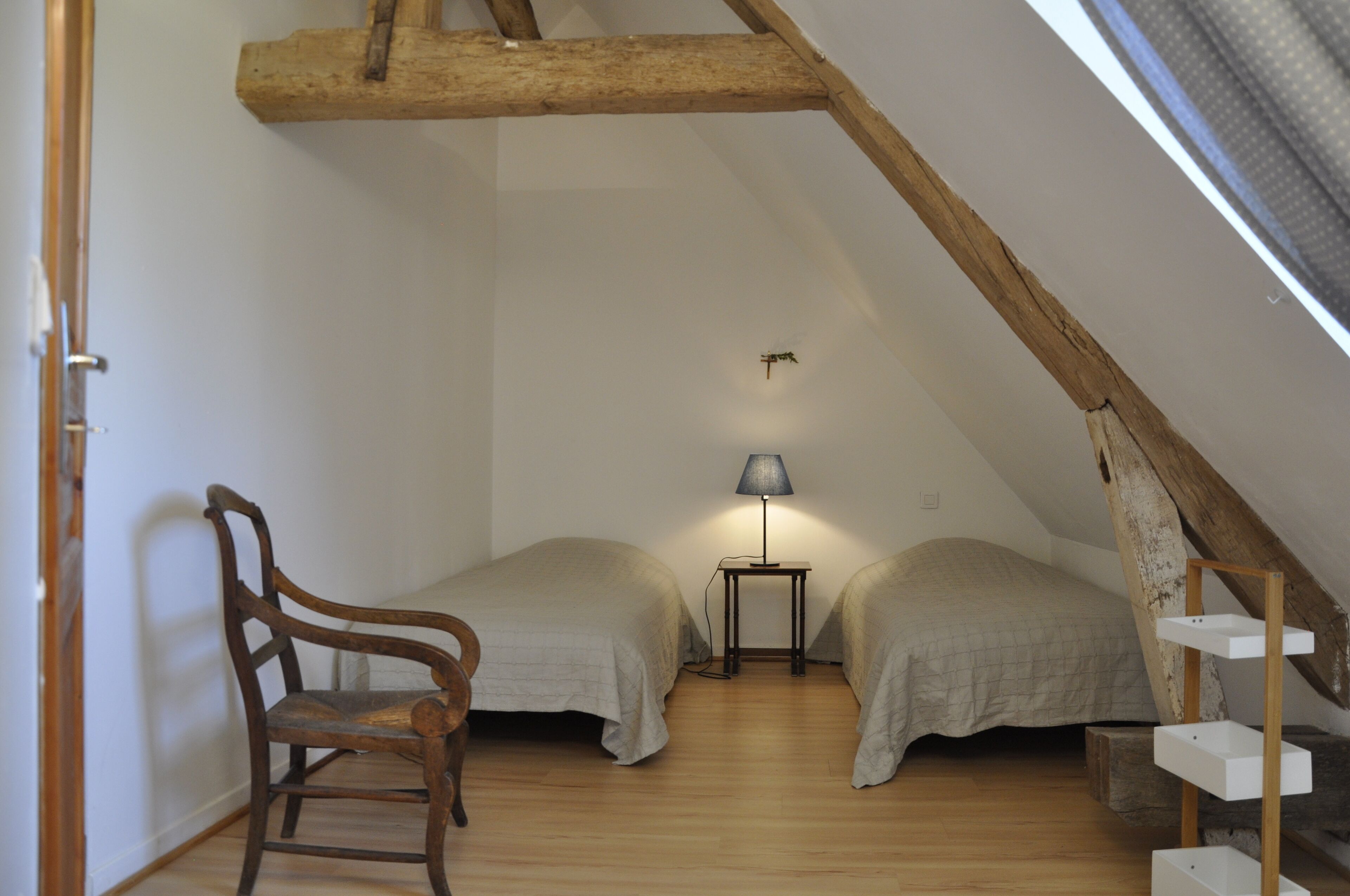 Un dormitorio abrigado y pequeño en el ático, con dos camas individuales, una silla de madera, una mesita con una lámpara y un techo inclinado.