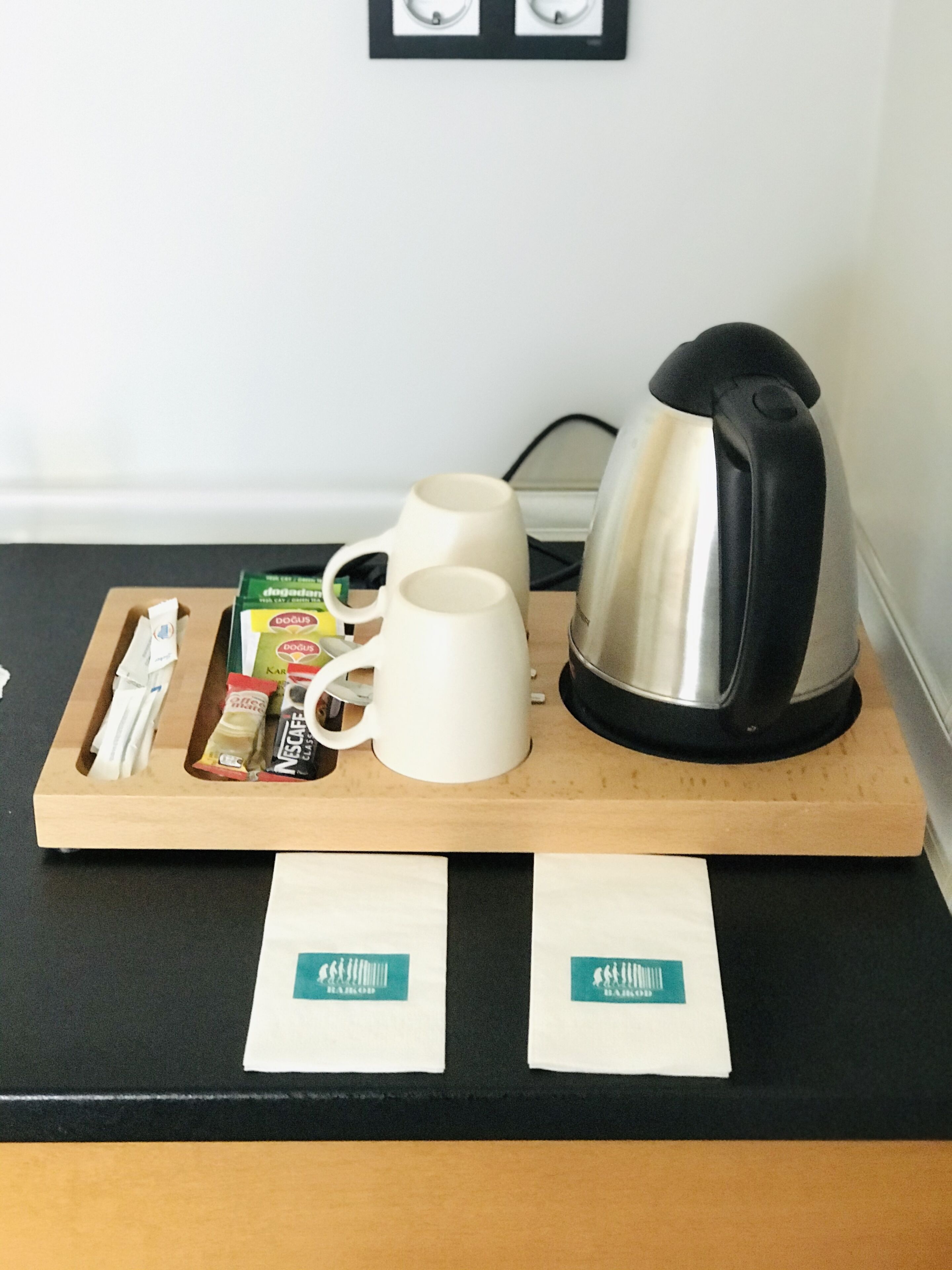 A wooden tray with a coffee maker, two white mugs, a box of coffee pods, and two sachets of coffee on a black countertop.
