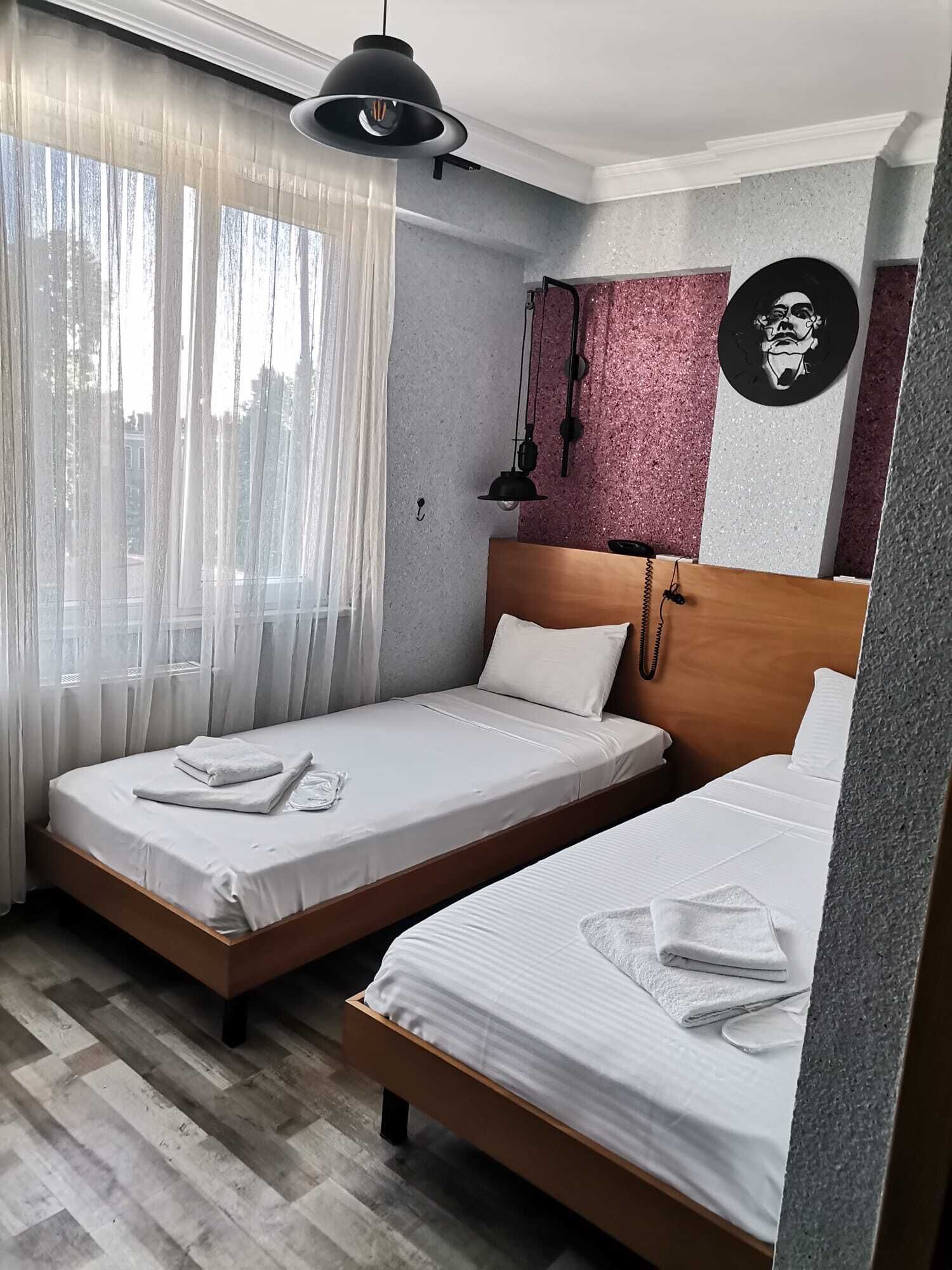 Habitació d'hotel amb dos llits, un gran finestral amb cortines transparents, una obra penjada a la paret i una làmpada cònica suspesa.